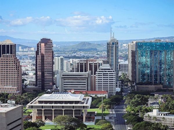 A cityscape featuring tall buildings, including modern skyscrapers, with mountains and a partly cloudy sky in the background.