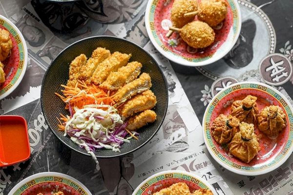 The image shows various plates of crispy fried food, including chicken and other items, arranged around a central dish on a table with a printed cloth.