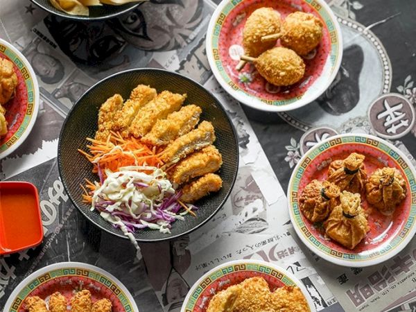The image shows various plates of crispy fried food, including chicken and other items, arranged around a central dish on a table with a printed cloth.