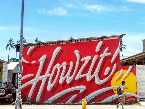 A mural with the text "Howzit!" in bold white letters on a red background, with a person walking by and palm trees in the background.
