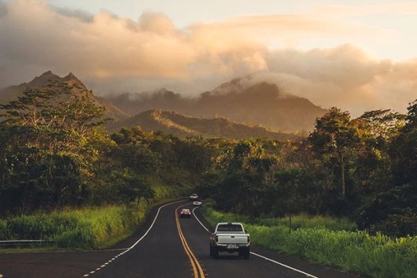 A scenic road with a vehicle driving through a lush, mountainous landscape surrounded by greenery and enveloped in soft, golden sunlight and clouds.