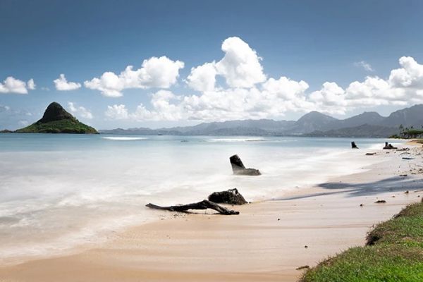 A serene beach scene with clear water, scattered driftwood on the sand, distant mountains, and a unique island formation under a partly cloudy sky.