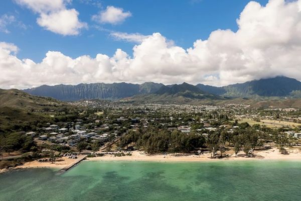 A coastal landscape with a sandy beach and turquoise waters, bordered by a residential area and mountains under a partly cloudy sky.