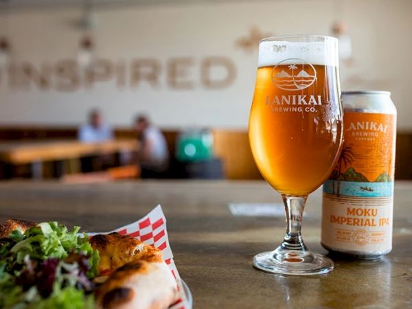 A glass of beer, a can labeled "Lanikai Brewing Co. Moku Imperial IPA," and a plate with food are on a table in a casual dining setting.