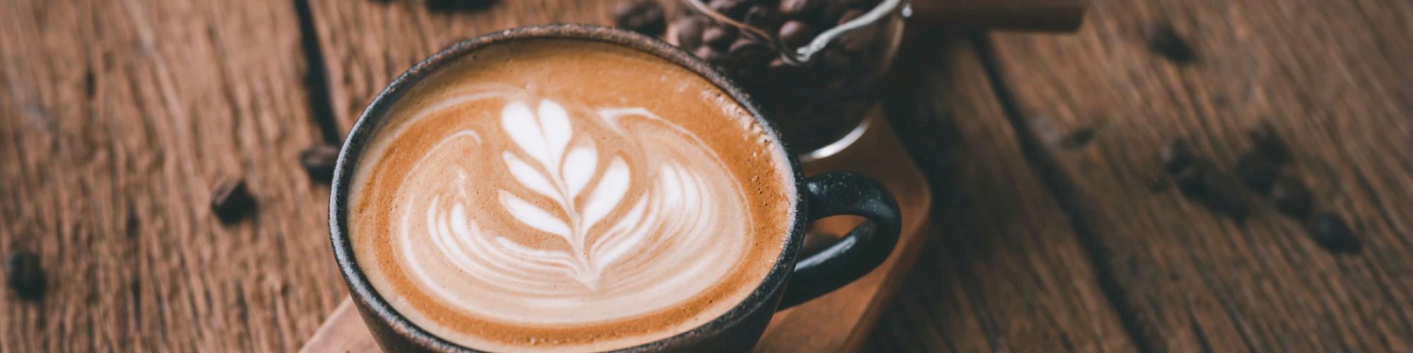 A latte with latte art in a ceramic cup sits on a wooden surface near a small glass container of coffee beans.