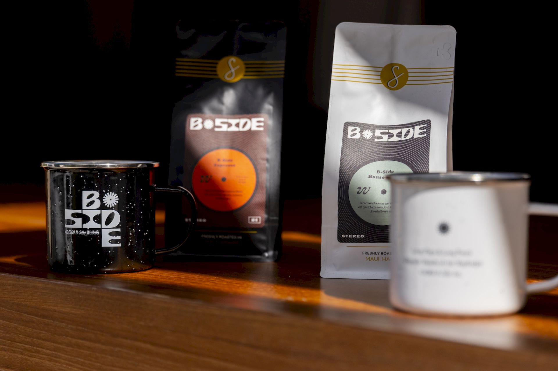 A wooden table with B-Side coffee packaging and a black mug labeled &ldquo;3rd Side,&rdquo; plus a blurred cup and spoon nearby.