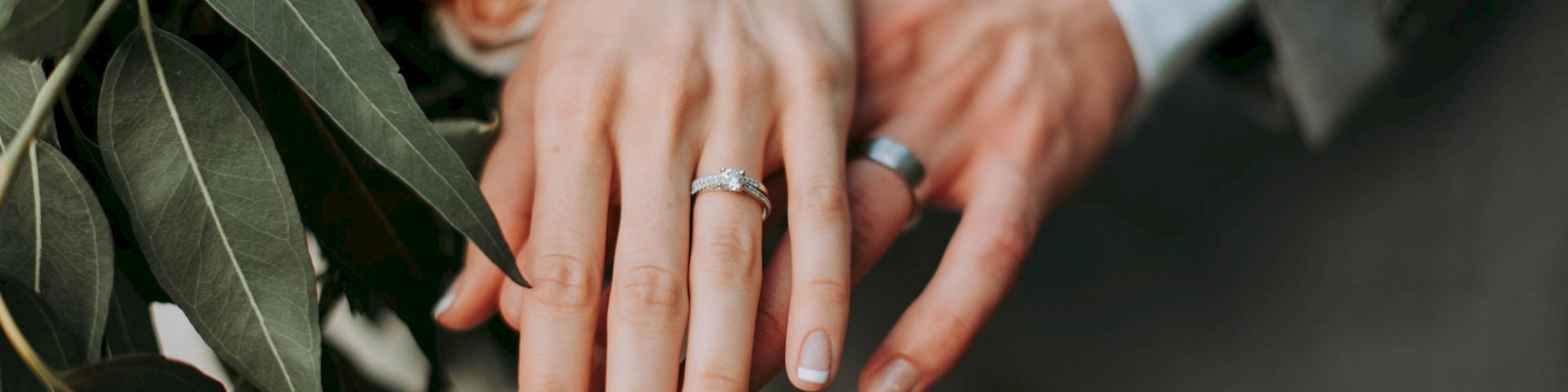 A close-up of two hands wearing wedding rings, layered over each other with a bouquet of flowers nearby. The background is slightly blurred.