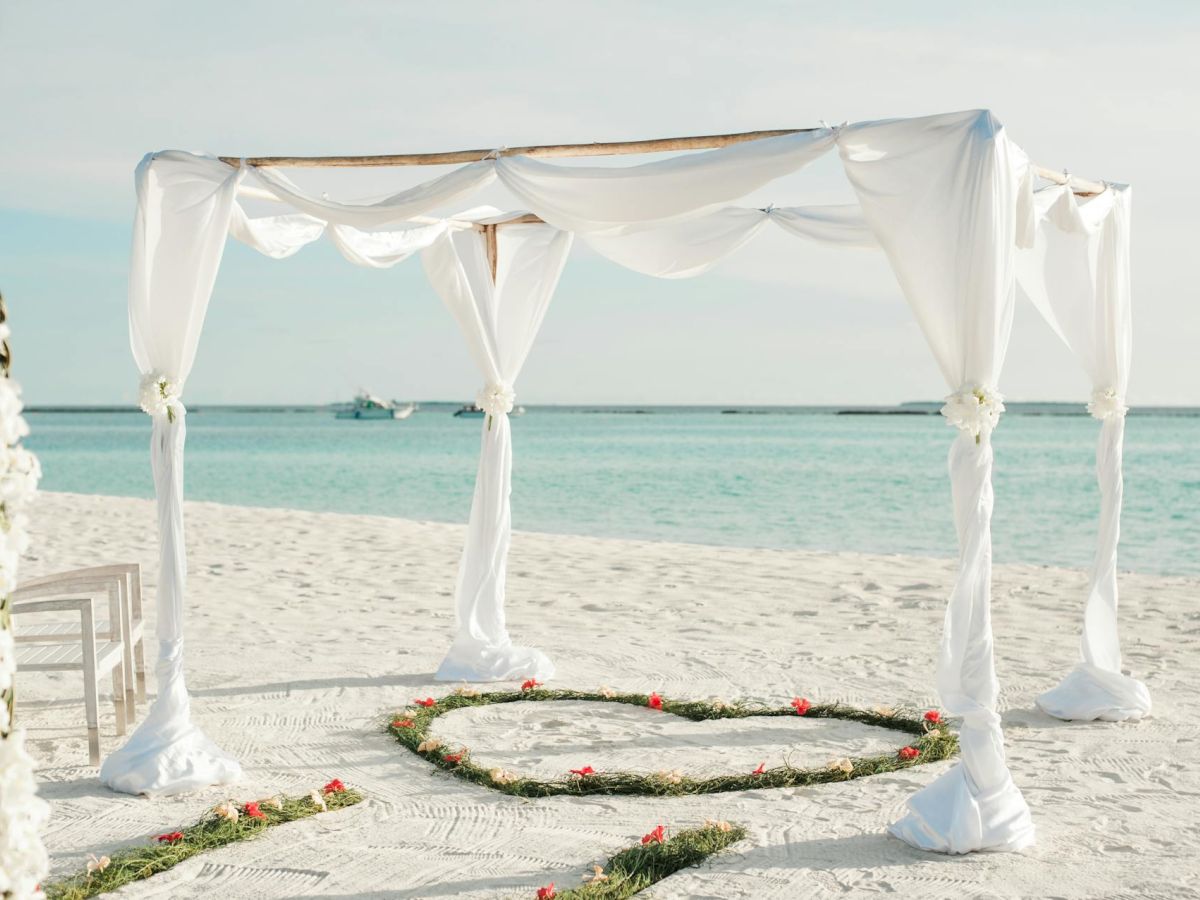 A beachside wedding altar with white drapes, a floral arch, and a heart shape on the sand is set up near the shore on a sunny day.