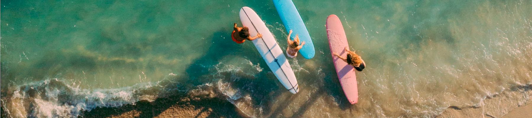 Three surfers holding surfboards—pink, blue, and white—stand at the shoreline, with clear turquoise waves gently washing over their feet.