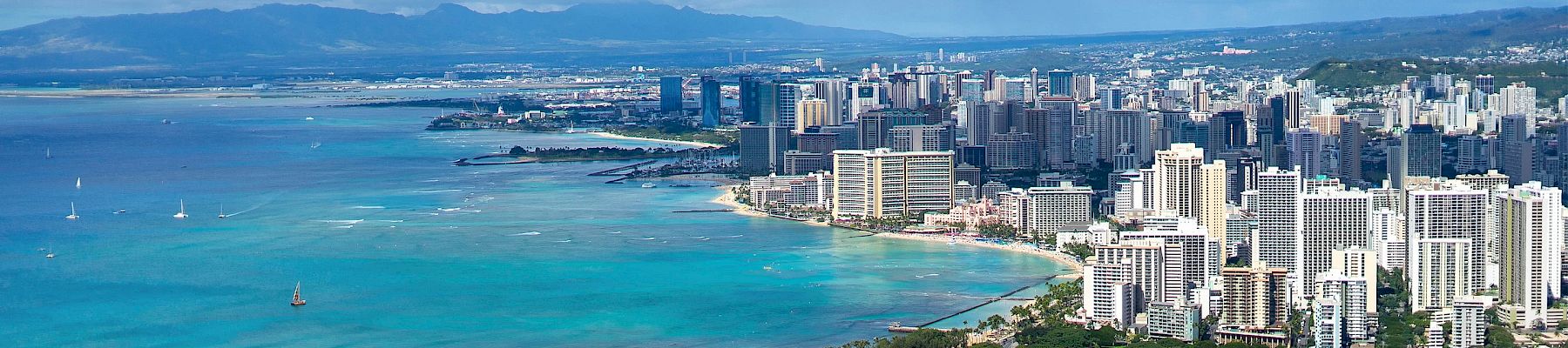 Aerial view of a coastal city with numerous skyscrapers, clear turquoise waters, boats in the water, and mountains in the background.