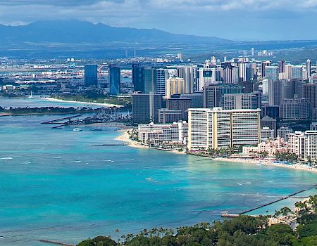 Aerial view of a coastal city with numerous skyscrapers, clear turquoise waters, boats in the water, and mountains in the background.