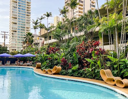 A beautiful outdoor pool area surrounded by lush greenery, modern buildings, and seating areas with umbrellas and lounge chairs along the edge.