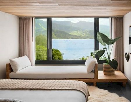 A serene bedroom features a window bench, a large plant, a wall tapestry, and a stunning view of mountains and water through large windows.