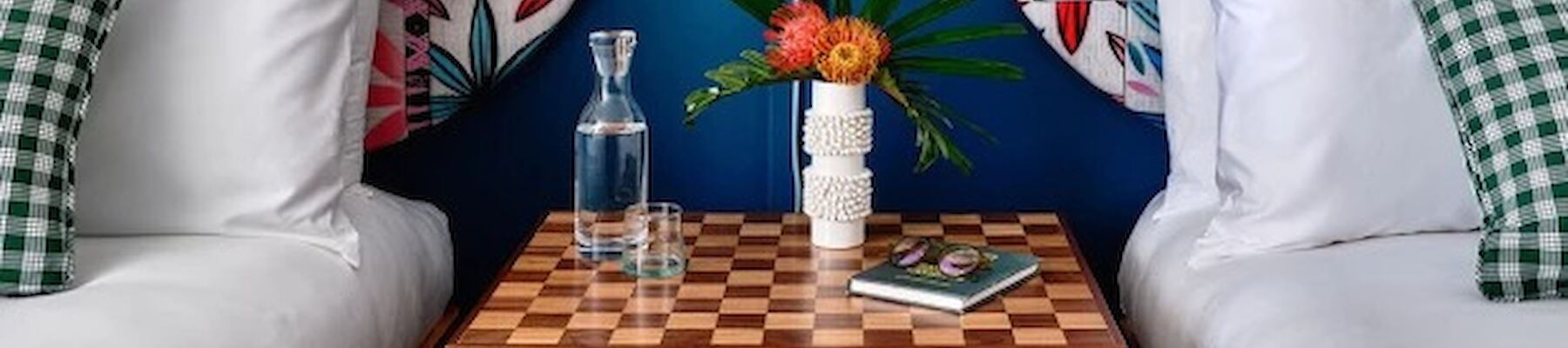 A stylish bedroom with two beds, patterned headboards, checkerboard nightstand, books, a vase with flowers, and a mid-century pendant light ending the sentence.