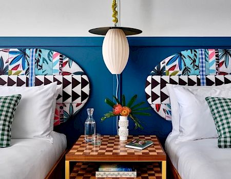 A stylish bedroom with two beds, patterned headboards, checkerboard nightstand, books, a vase with flowers, and a mid-century pendant light ending the sentence.