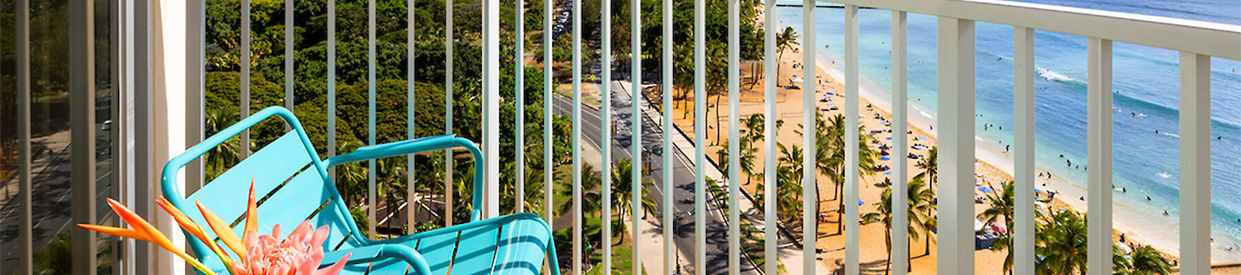 A balcony view overlooks a beach with clear blue waters, showing a table with a flower and a cup, beside two chairs, and a mountain in the distance.