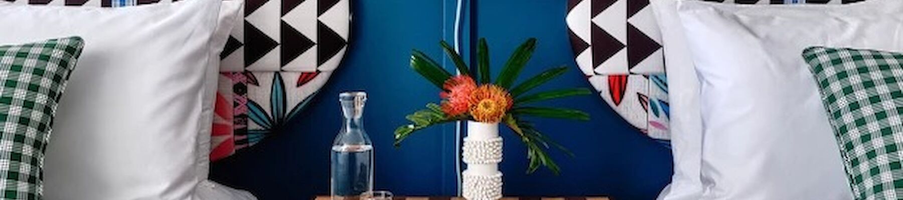 A stylish bedroom features two beds with vibrant headboards, a central hanging lamp, a checkered bedside table with books, flowers, and a water carafe.