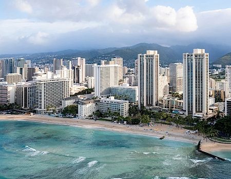 A beautiful coastal cityscape with tall buildings, a sandy beach, and a calm sea, set against a backdrop of mountains and a partly cloudy sky.