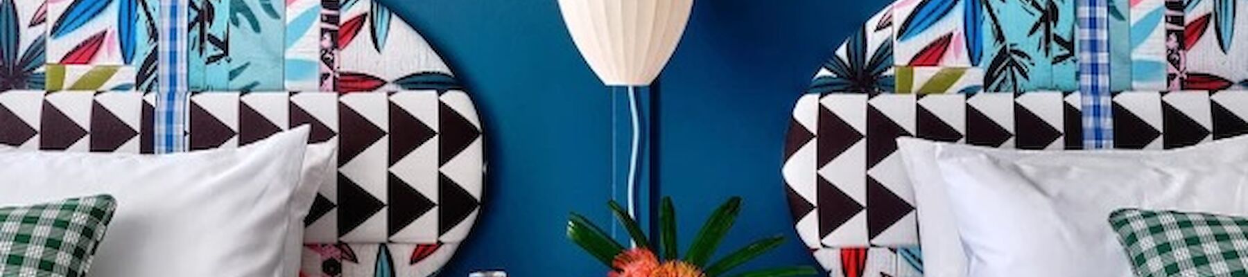 A bedroom with two beds, colorful headboards, checkered bedside table, decorative pillows, and a pendant light above. White and blue theme.