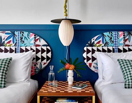 A bedroom with two beds, colorful headboards, checkered bedside table, decorative pillows, and a pendant light above. White and blue theme.