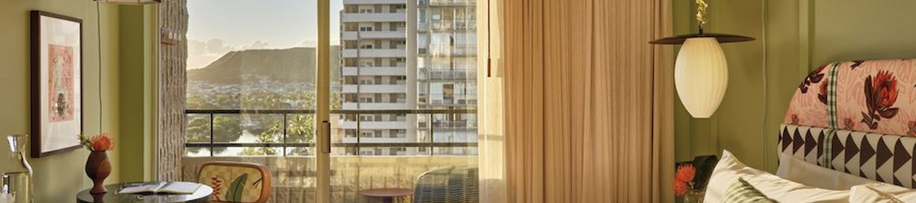 A cozy, well-lit room with a bed, green walls, a small dining area, and a balcony overlooking city buildings and mountains in the background.