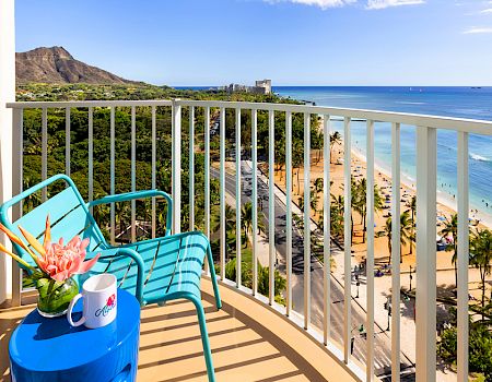 A balcony with blue chairs and a table overlooks a scenic beach and ocean, with mountains in the background and clear blue skies above.