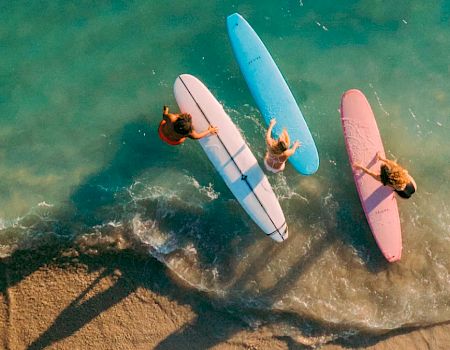 Three surfers with orange, blue, and pink surfboards are in shallow turquoise waters near the sandy shore, casting long shadows.