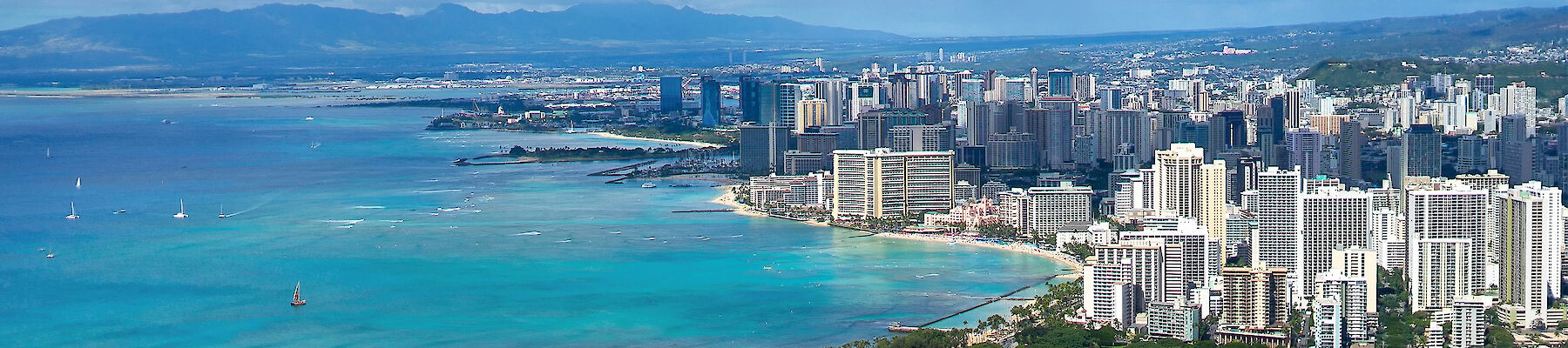 A coastal cityscape with numerous high-rise buildings, boats on turquoise waters, and mountains in the background under a partly cloudy sky.