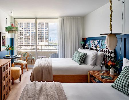 A modern hotel room features two neatly made beds, a balcony with a city view, and a stylish decor with checkered pillows and artistic lighting.