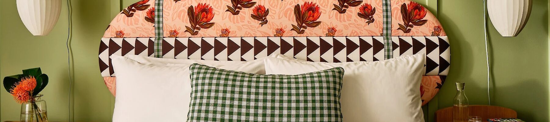 A neatly made bed features a floral-patterned headboard and gingham pillow, flanked by hanging pendant lights and nightstands with small decor items.