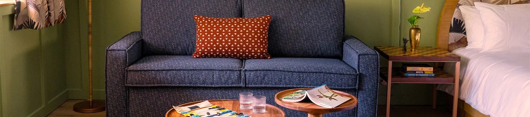 A cozy living space with a blue sofa, framed art, wooden coffee tables, a bed, and a lamp. Magazines and a flower vase on the tables.