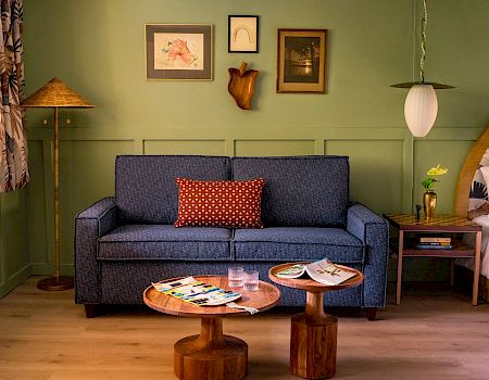A cozy living space with a blue sofa, framed art, wooden coffee tables, a bed, and a lamp. Magazines and a flower vase on the tables.
