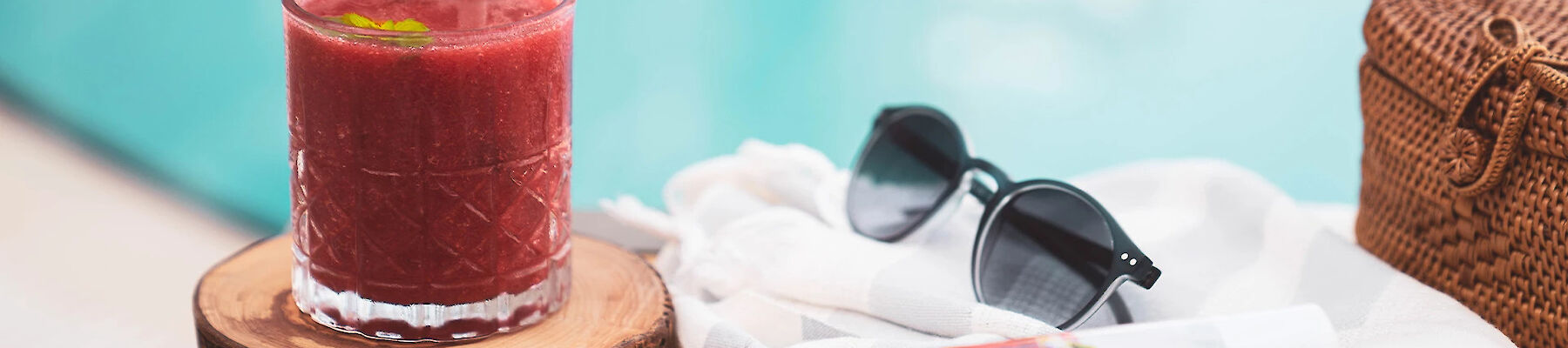 A glass of red beverage with mint, sunglasses, a woven bag, and a rolled towel with a magazine beside a pool.