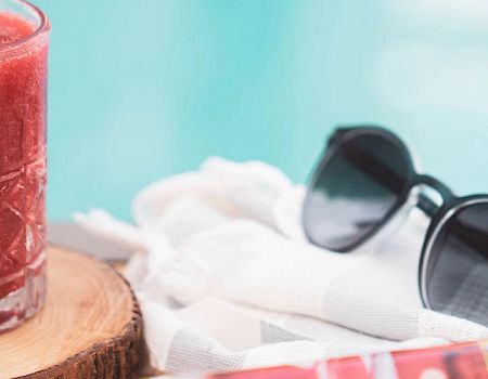 A glass of red beverage with mint, sunglasses, a woven bag, and a rolled towel with a magazine beside a pool.