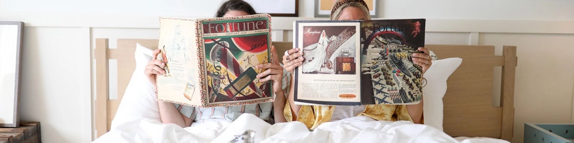 Two people in bed are reading magazines, mostly covering their faces. The room has framed pictures on the wall and a tidy appearance.