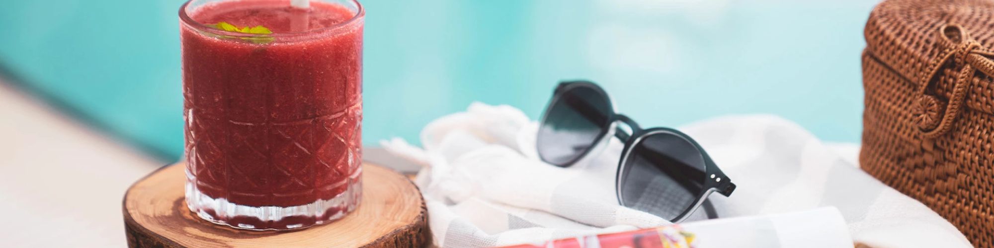 The image shows a red drink in a glass, sunglasses, a woven bag, and a rolled-up magazine or towel placed beside a pool.