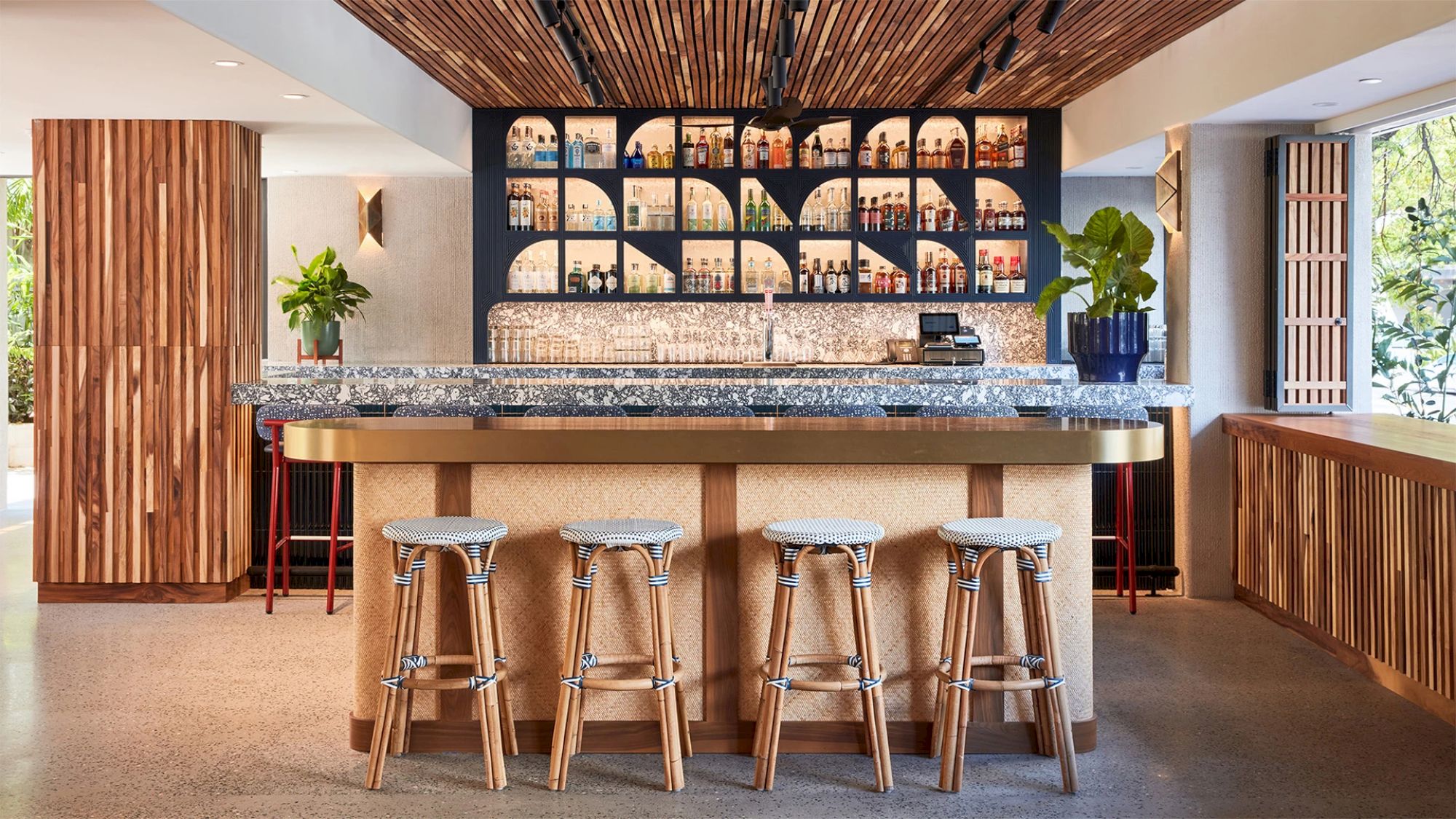 A modern bar area with four wooden stools, a marble counter, plants, and an illuminated wall displaying various bottles.