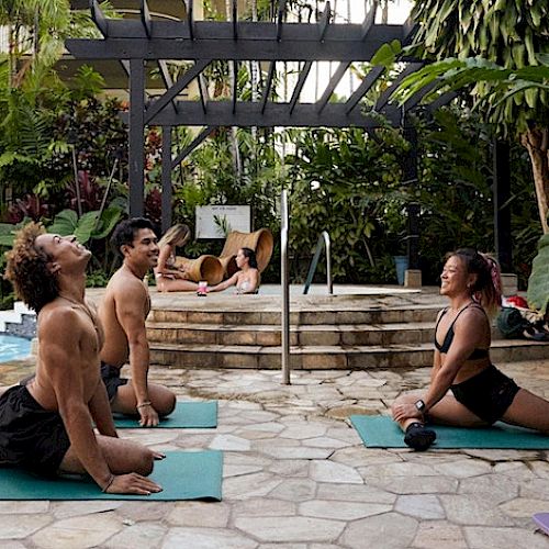 People practice yoga on mats by a pool, with lush greenery in the background.