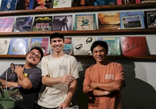 Three people are standing in front of a wall displaying various vinyl record covers on wooden shelves. They appear to be smiling.