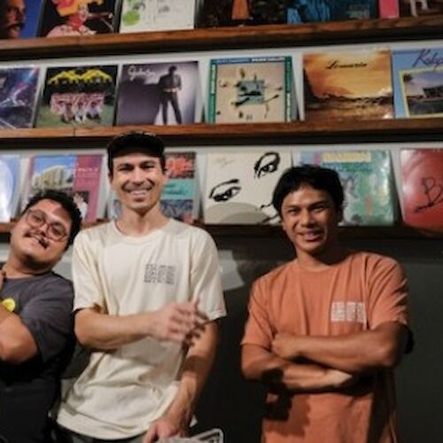 Three people are standing in front of a wall displaying various vinyl record covers on wooden shelves. They appear to be smiling.