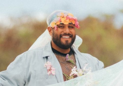 A person is outdoors, smiling and wearing a crown of flowers on their head with a sheer veil. The background is blurry and natural.