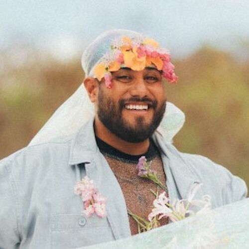 A person is outdoors, smiling and wearing a crown of flowers on their head with a sheer veil. The background is blurry and natural.