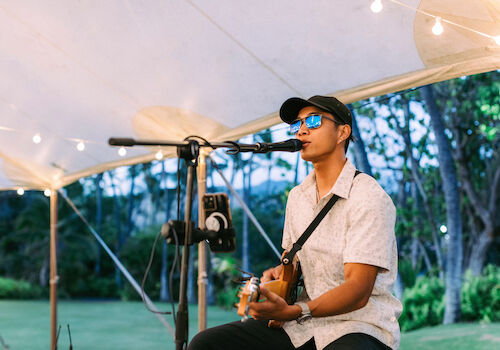 A person wearing sunglasses and a hat sits under a tent, playing an electric guitar and singing into a microphone.