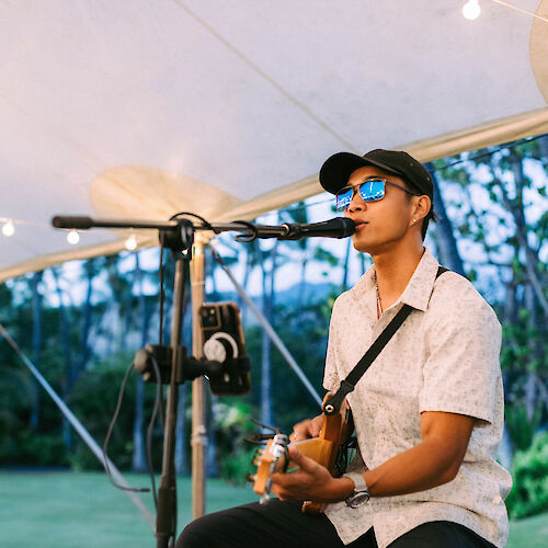 A person wearing sunglasses and a hat sits under a tent, playing an electric guitar and singing into a microphone.