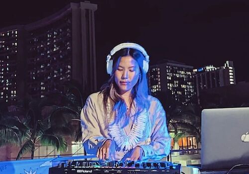 A person is DJing outdoors at night, wearing headphones with a mixing console in front and a laptop nearby. Tall buildings are in the background.