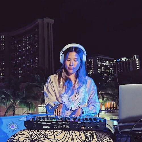 A person is DJing outdoors at night, wearing headphones with a mixing console in front and a laptop nearby. Tall buildings are in the background.