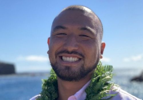 A person smiles broadly at the camera against a backdrop of the ocean and blue sky, while wearing a green lei around their neck.