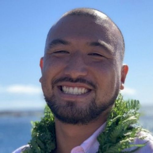 A person smiles broadly at the camera against a backdrop of the ocean and blue sky, while wearing a green lei around their neck.