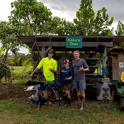 People pose at a lush roadside coffee stand under big trees with a green sign reading &ldquo;Kāko&rsquo;o ʻOiwi.&rdquo;