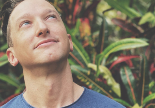 A person in a blue shirt looks upwards while standing in front of colorful foliage in a natural setting.